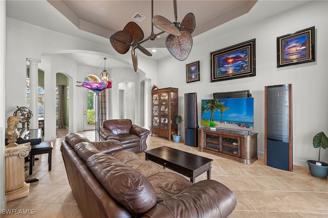 tiled living room featuring decorative columns, a tray ceiling, and ceiling fan with notable chandelier