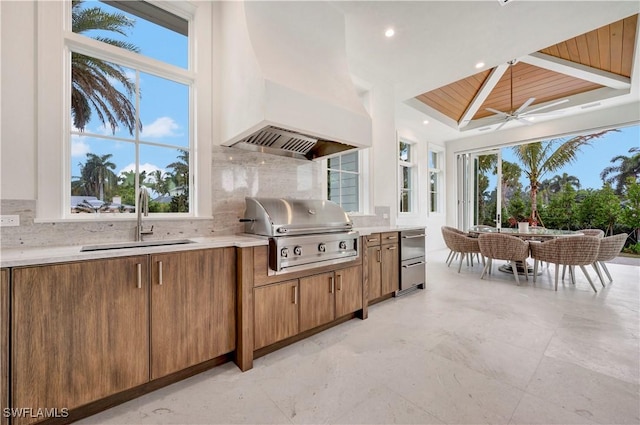 kitchen with brown cabinetry, custom range hood, backsplash, light countertops, and a sink