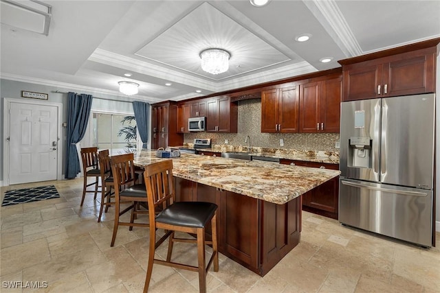 kitchen with decorative backsplash, appliances with stainless steel finishes, a tray ceiling, a kitchen island, and light stone counters