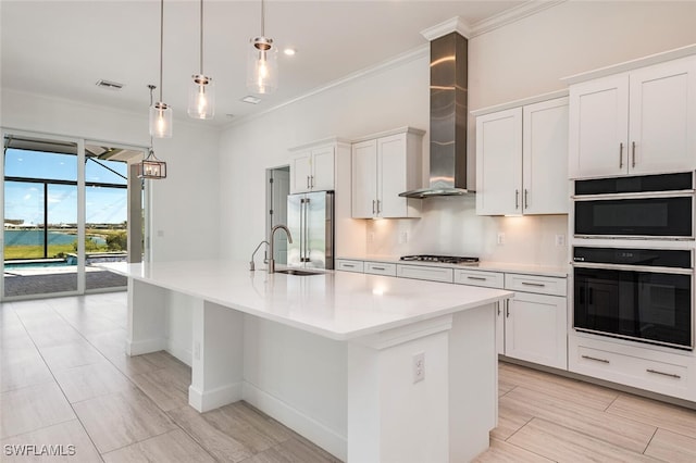 kitchen featuring pendant lighting, a center island with sink, wall chimney range hood, white cabinetry, and stainless steel appliances