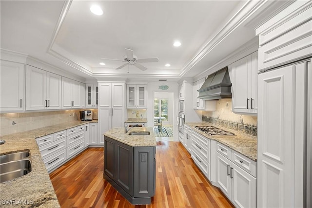 kitchen featuring custom exhaust hood, a tray ceiling, sink, a center island with sink, and white cabinetry