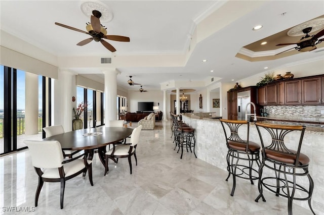 dining space with ornate columns, ceiling fan with notable chandelier, sink, a tray ceiling, and crown molding