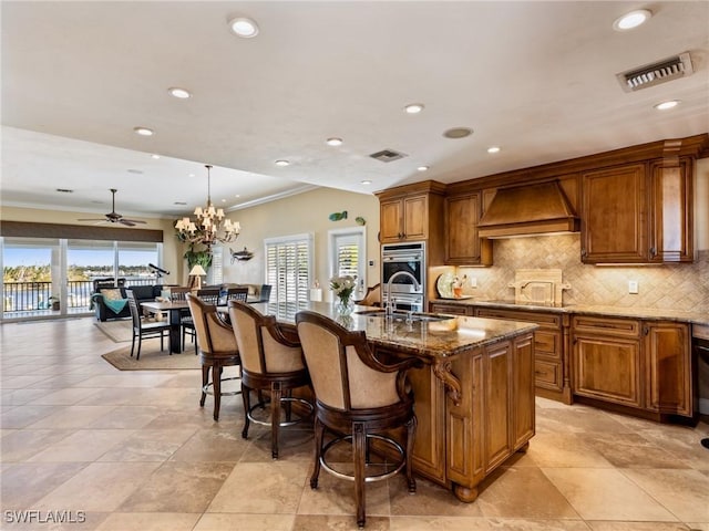 kitchen with hanging light fixtures, custom range hood, dark stone countertops, a center island with sink, and ceiling fan with notable chandelier