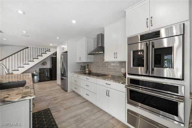 kitchen with appliances with stainless steel finishes, white cabinetry, a sink, wall chimney range hood, and light stone countertops