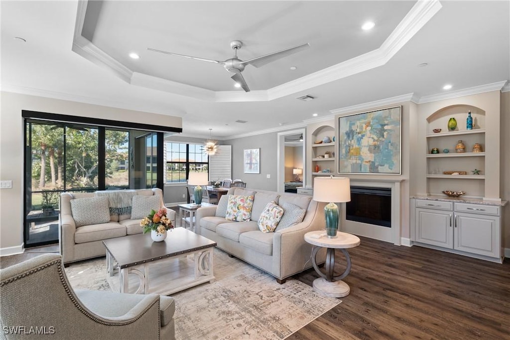 living room with ornamental molding, ceiling fan with notable chandelier, a tray ceiling, hardwood / wood-style flooring, and built in features