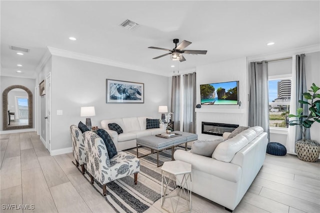 living area featuring light wood finished floors, ornamental molding, a glass covered fireplace, and visible vents