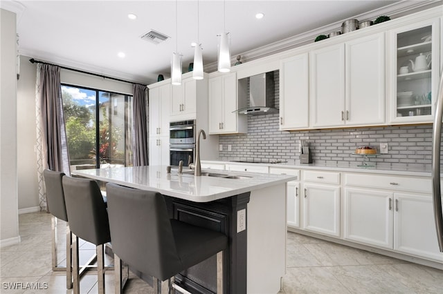 kitchen with pendant lighting, wall chimney exhaust hood, an island with sink, white cabinetry, and stainless steel double oven