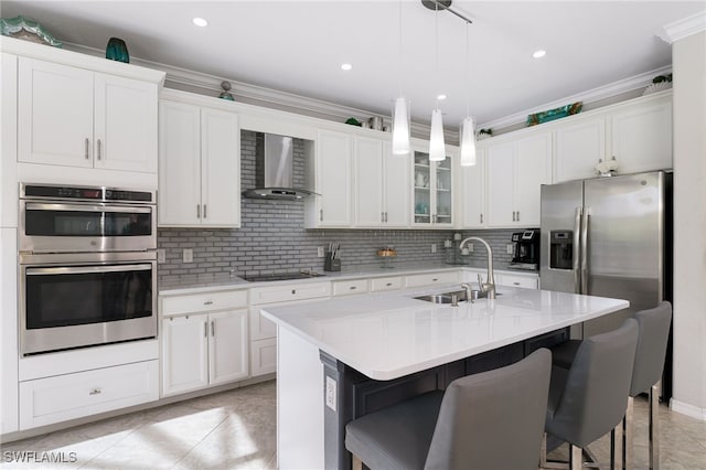 kitchen with sink, wall chimney range hood, an island with sink, appliances with stainless steel finishes, and ornamental molding