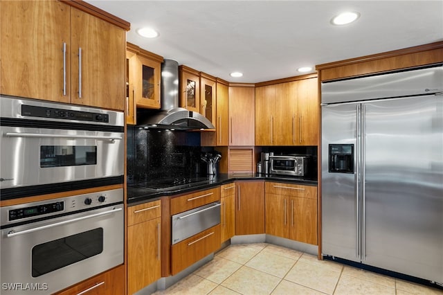 kitchen featuring wall chimney exhaust hood, stainless steel appliances, backsplash, dark stone counters, and light tile patterned flooring