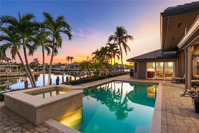 pool at dusk with an in ground hot tub, a patio, and a water view