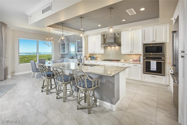 kitchen featuring stainless steel appliances, wall chimney range hood, pendant lighting, white cabinets, and an island with sink