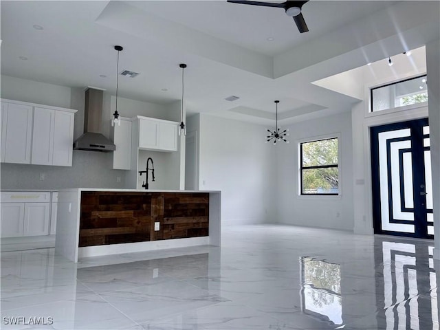 kitchen with pendant lighting, ceiling fan with notable chandelier, a raised ceiling, wall chimney range hood, and white cabinetry