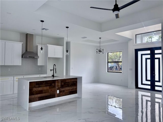 kitchen with a tray ceiling, white cabinetry, a kitchen island with sink, and wall chimney range hood