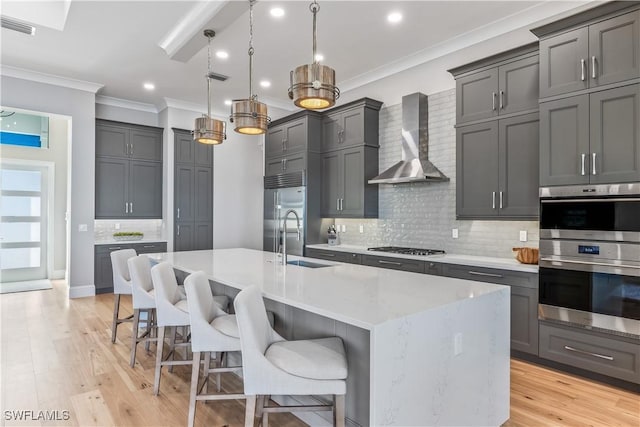 kitchen featuring a breakfast bar area, hanging light fixtures, stainless steel appliances, an island with sink, and wall chimney exhaust hood