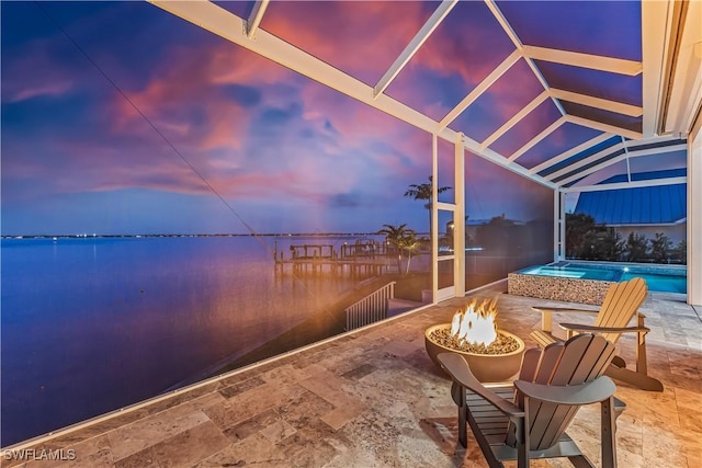 patio terrace at dusk featuring a water view, glass enclosure, and a fire pit