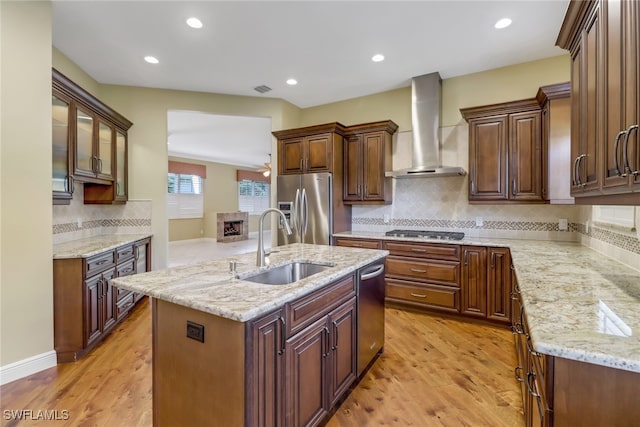 kitchen with light wood-style flooring, stainless steel appliances, a sink, wall chimney exhaust hood, and an island with sink