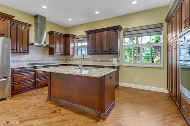 kitchen featuring a center island with sink, appliances with stainless steel finishes, light stone counters, wall chimney range hood, and a sink