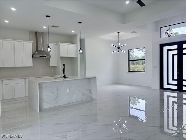 kitchen featuring sink, wall chimney exhaust hood, a notable chandelier, a kitchen island with sink, and white cabinets