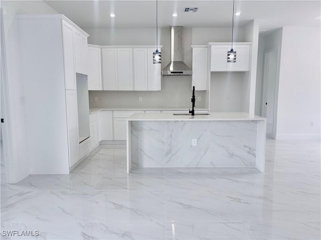 kitchen with sink, wall chimney exhaust hood, an island with sink, pendant lighting, and white cabinets