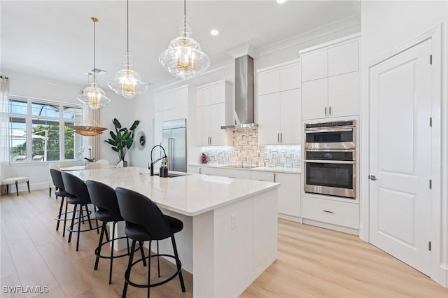 kitchen with appliances with stainless steel finishes, wall chimney range hood, pendant lighting, a center island with sink, and white cabinets