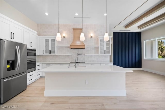 kitchen with hanging light fixtures, appliances with stainless steel finishes, a kitchen island with sink, white cabinets, and custom exhaust hood