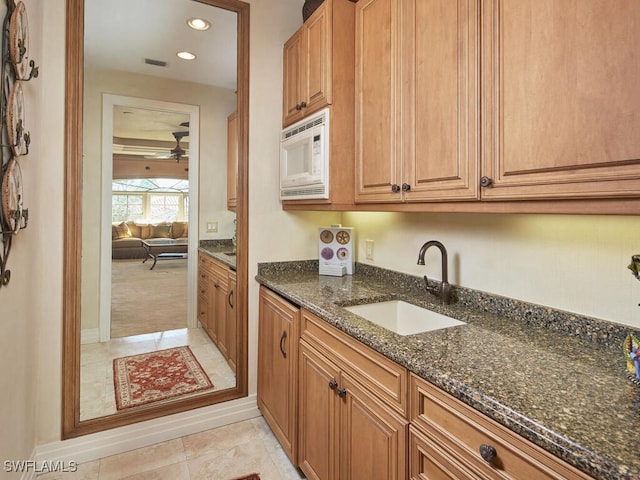 kitchen with light tile patterned floors, white microwave, brown cabinetry, a sink, and dark stone counters