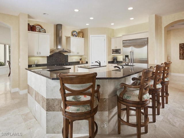 kitchen featuring stainless steel appliances, wall chimney range hood, a large island with sink, and white cabinets