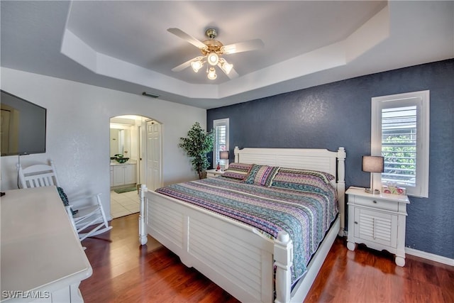 bedroom with ensuite bathroom, ceiling fan, dark wood-type flooring, and a tray ceiling