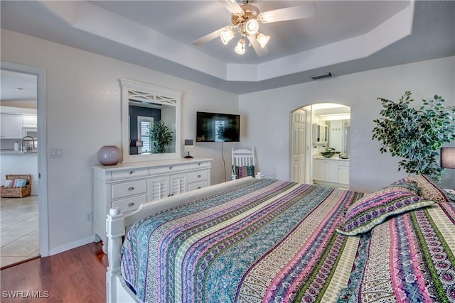 bedroom featuring a raised ceiling, ensuite bath, ceiling fan, and light wood-type flooring