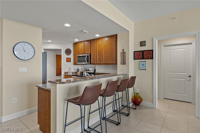 kitchen featuring kitchen peninsula, appliances with stainless steel finishes, dark stone counters, a breakfast bar, and light tile patterned floors