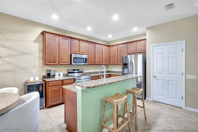 kitchen with stainless steel appliances, wine cooler, light stone counters, a kitchen bar, and a kitchen island with sink