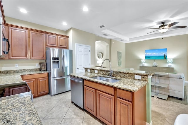 kitchen with a center island with sink, sink, ceiling fan, light stone counters, and stainless steel appliances