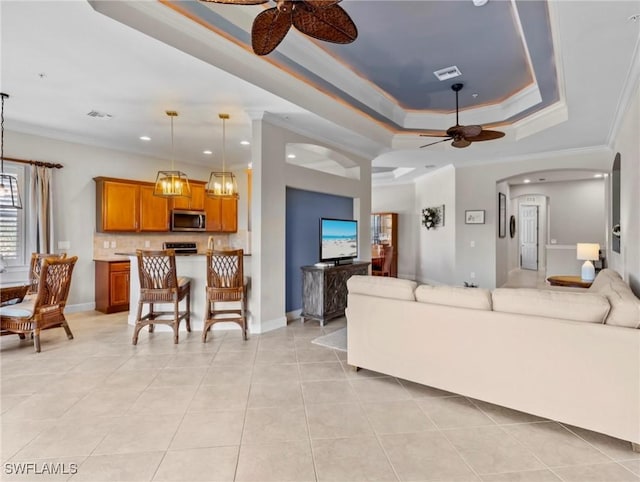 living room with ceiling fan with notable chandelier, a raised ceiling, ornamental molding, and light tile patterned floors