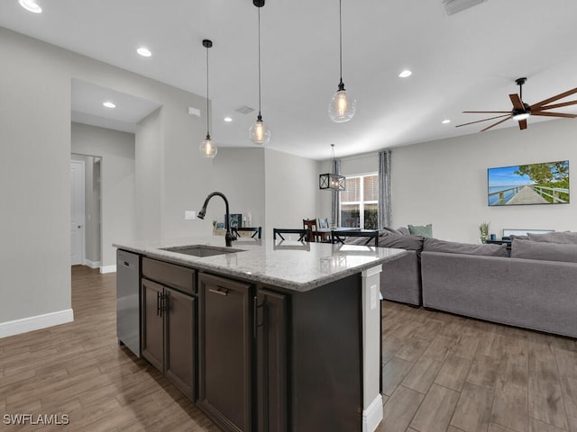 kitchen featuring a kitchen island with sink, a sink, open floor plan, stainless steel dishwasher, and light stone countertops