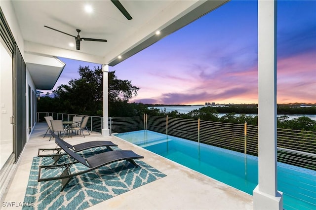 pool at dusk featuring a water view, outdoor dining space, a ceiling fan, and fence