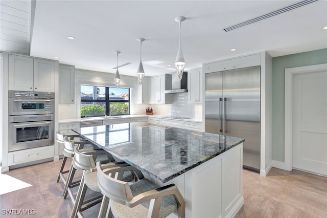 kitchen with dark stone counters, wall chimney exhaust hood, stainless steel appliances, pendant lighting, and a kitchen island