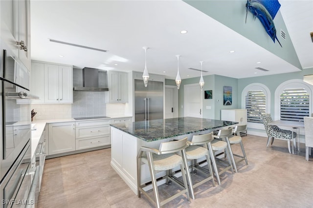 kitchen featuring built in fridge, wall chimney range hood, a kitchen island, a kitchen bar, and white cabinetry