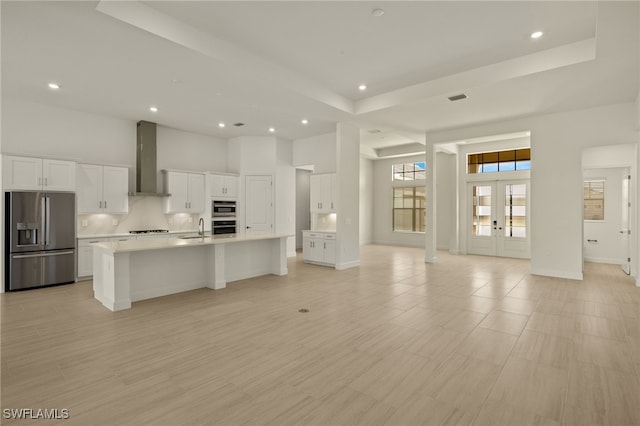 kitchen with sink, wall chimney exhaust hood, a kitchen island with sink, white cabinets, and appliances with stainless steel finishes