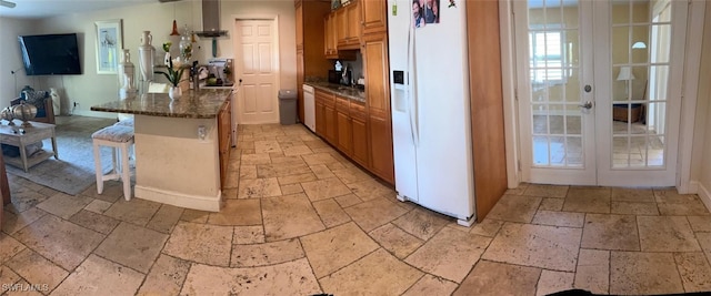 kitchen with white appliances, dark stone counters, french doors, a kitchen breakfast bar, and wall chimney exhaust hood