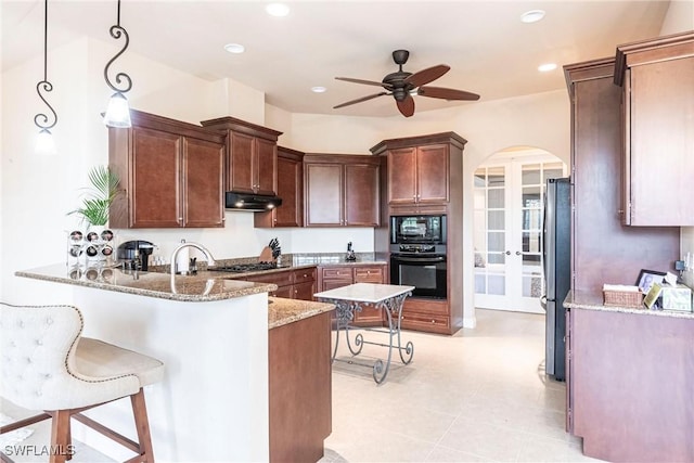 kitchen with arched walkways, under cabinet range hood, hanging light fixtures, light stone countertops, and black appliances