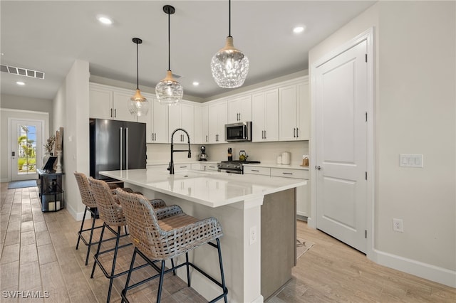 kitchen with appliances with stainless steel finishes, light wood-type flooring, pendant lighting, white cabinetry, and an island with sink