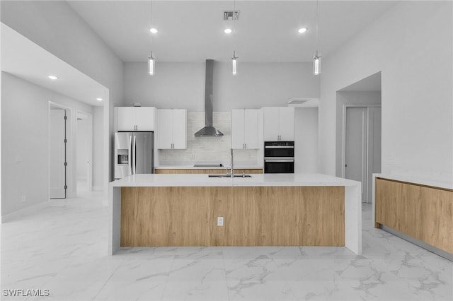 kitchen featuring wall chimney exhaust hood, decorative light fixtures, stainless steel fridge with ice dispenser, white cabinetry, and an island with sink