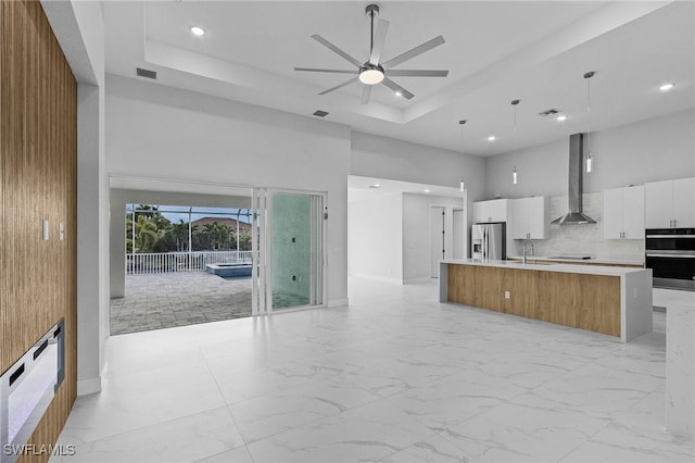 kitchen with stainless steel refrigerator with ice dispenser, a raised ceiling, wall chimney range hood, a large island with sink, and white cabinetry