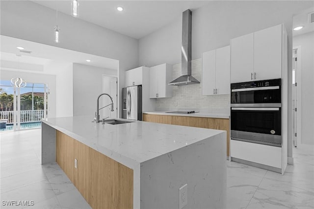 kitchen featuring white cabinetry, wall chimney exhaust hood, stainless steel appliances, hanging light fixtures, and an island with sink