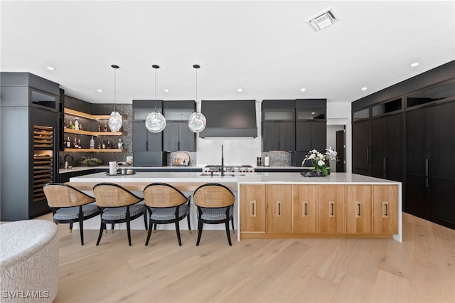 kitchen with premium range hood, a kitchen island with sink, decorative backsplash, decorative light fixtures, and light hardwood / wood-style floors