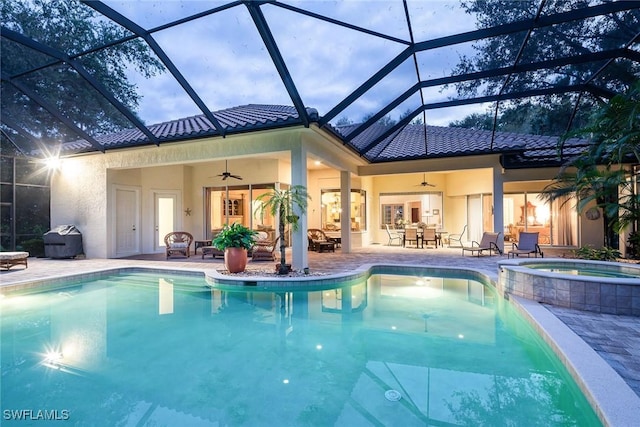 pool at dusk featuring glass enclosure, ceiling fan, a patio, and an in ground hot tub