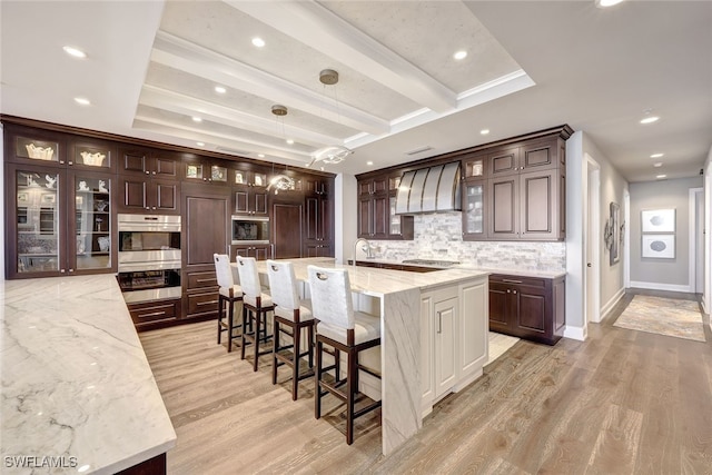 kitchen with light stone countertops, light hardwood / wood-style flooring, premium range hood, a large island with sink, and decorative light fixtures