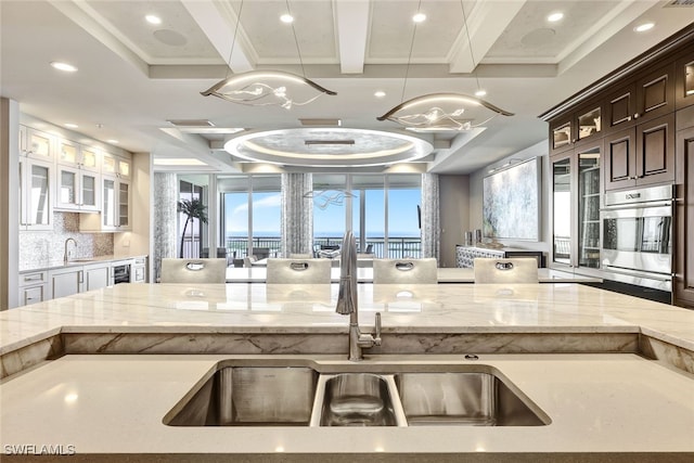 kitchen with stainless steel oven, tasteful backsplash, beamed ceiling, decorative light fixtures, and light stone counters