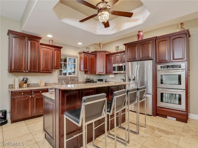 kitchen with a kitchen bar, appliances with stainless steel finishes, a raised ceiling, ceiling fan, and a center island