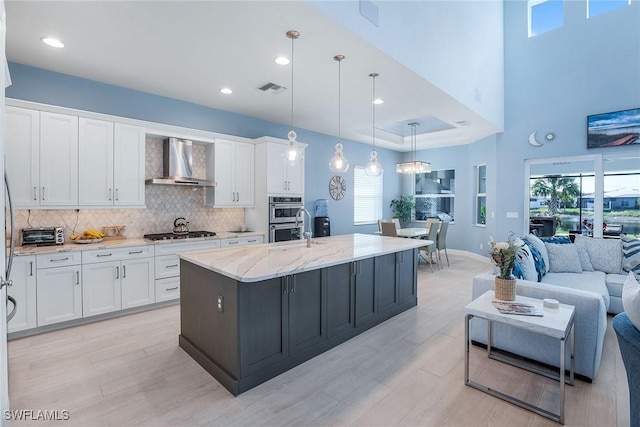 kitchen with white cabinets, an island with sink, decorative light fixtures, and wall chimney range hood